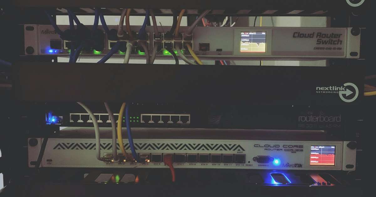 MikroTik en un rack de red: un Cloud Router Switch, un RouterBOARD RB3011 y un Cloud Core Router en funcionamiento, con cables y pantallas de tráfico
