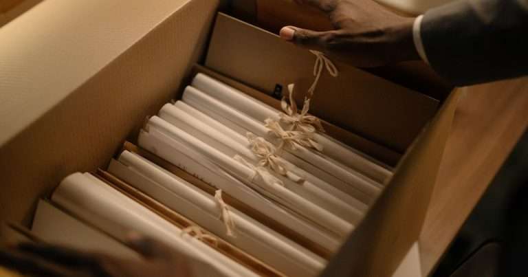 Archivar en Outlook: dos manos guardan en una caja de archivo de cartón una fila de legajos de papel blanco atados con cintas de algodón, como el correo antiguo que se saca del buzón para liberar espacio