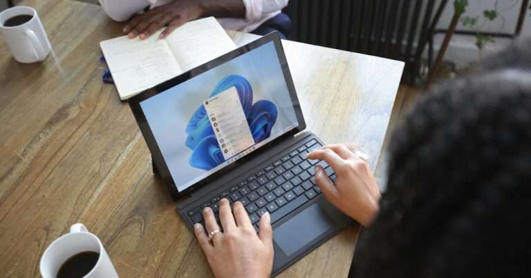 Buzón compartido en Outlook: dos personas trabajan en la misma mesa; una escribe en un portátil con Windows 11 y una lista de mensajes en pantalla, y la otra toma notas en un cuaderno