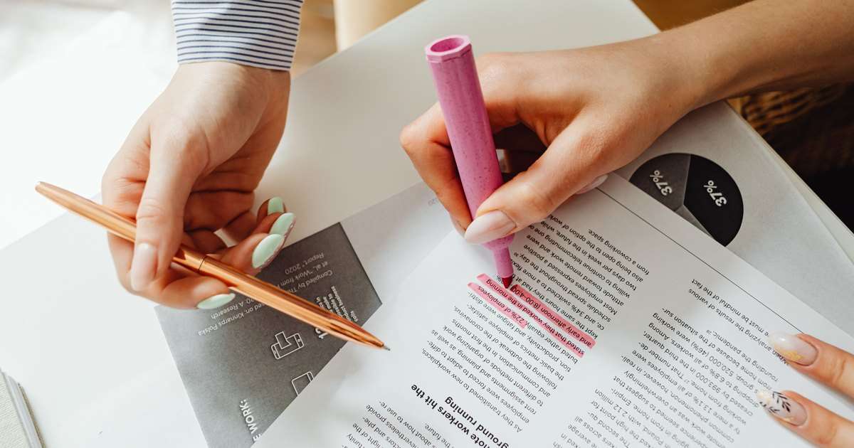 Control de cambios en Word: dos manos revisan un informe impreso sobre una mesa blanca; una resalta en rosa un párrafo con un rotulador y la otra sostiene un bolígrafo dorado, como las marcas que deja cada revisor