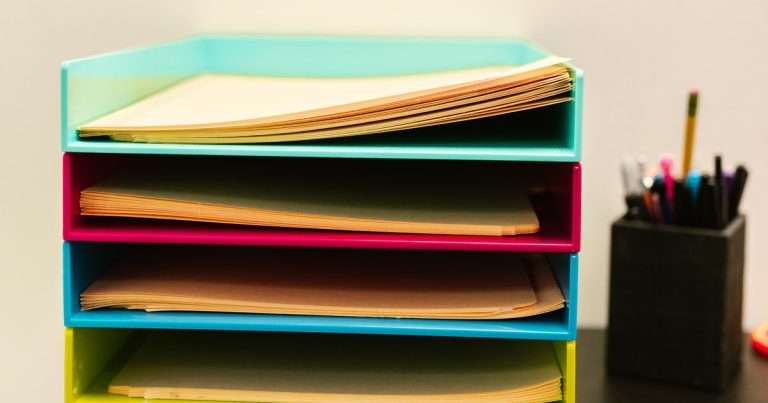 Filtros en Gmail: bandejas de documentos apiladas de colores turquesa, rosado, azul y verde, cada una con sus papeles, junto a un portalápices sobre un escritorio de oficina