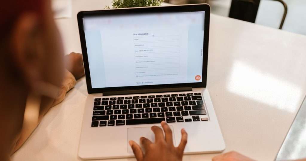 Formularios de Google: portátil sobre una mesa blanca con un formulario en línea de campos para nombre, dirección, fecha y correo, y una mano en el panel táctil