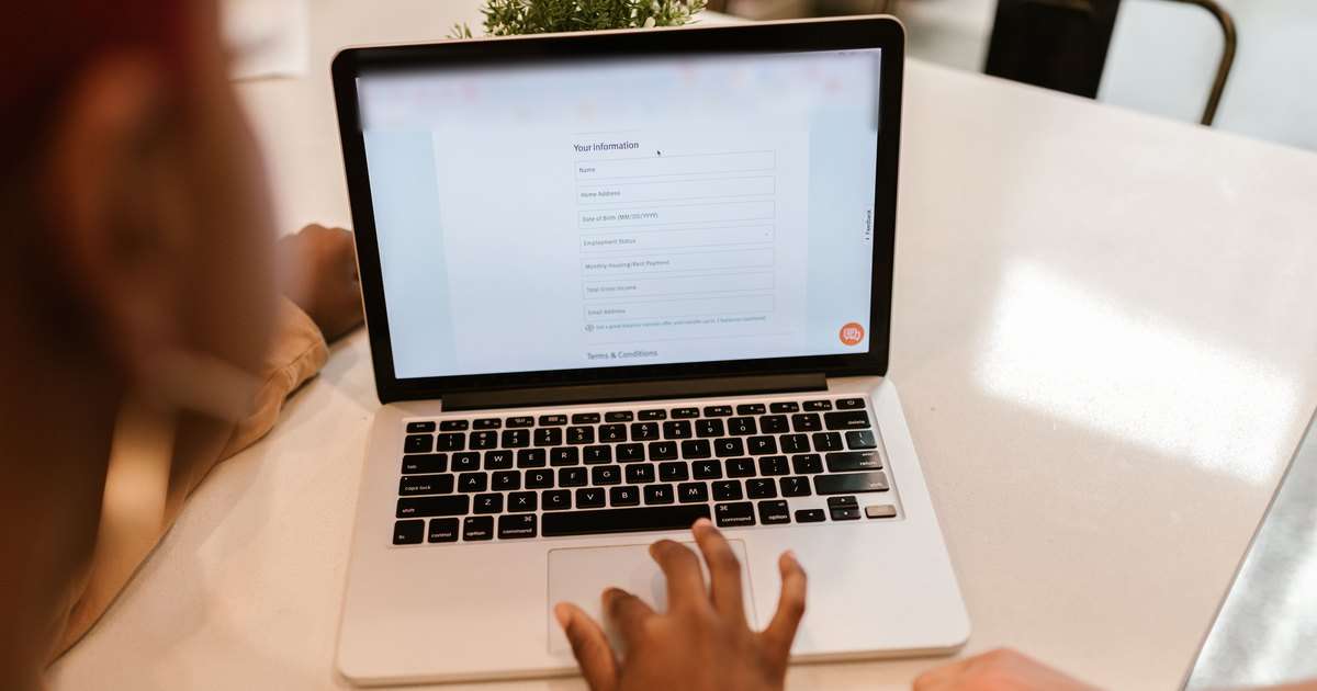 Formularios de Google: portátil sobre una mesa blanca con un formulario en línea de campos para nombre, dirección, fecha y correo, y una mano en el panel táctil