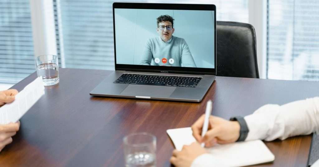 Grabar una reunión en Teams: un portátil sobre una mesa de juntas muestra a un participante remoto en videollamada mientras dos personas, de las que solo se ven las manos, leen un documento y toman notas