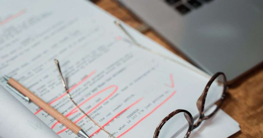 Historial de versiones: hoja impresa de un borrador con correcciones en rojo, un lápiz y unas gafas, junto a un portátil abierto sobre un escritorio de madera