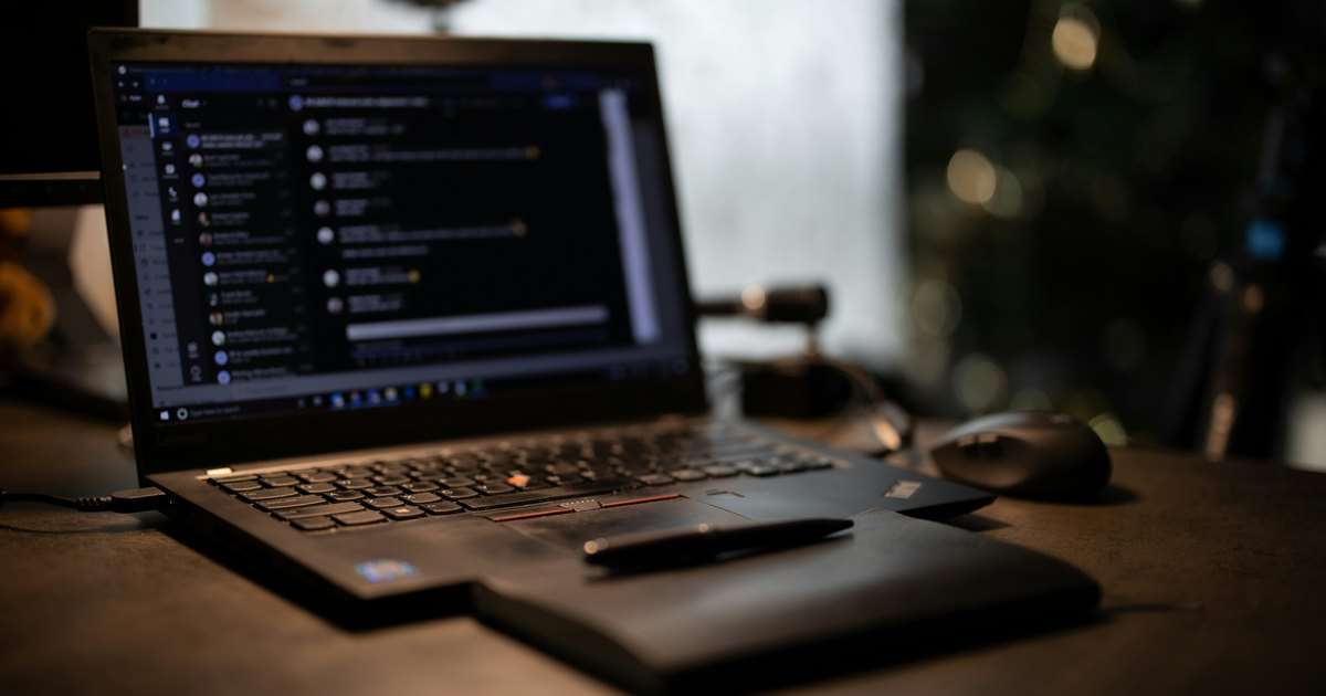 Microsoft Teams PowerShell administra lo que se ve aquí: un portátil con el chat de Microsoft Teams abierto en tema oscuro, sobre un escritorio con un ratón y un bolígrafo