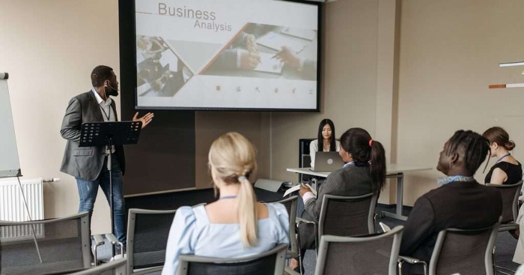 PowerPoint avanzado: un expositor, de pie junto a un atril, explica una diapositiva proyectada en la pantalla de una sala mientras varias personas lo escuchan sentadas