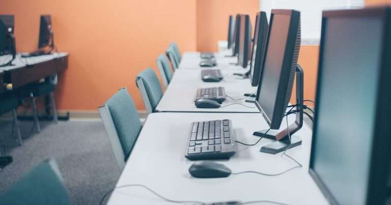 PowerShell remoto ejecuta con Invoke-Command la misma orden en todos los equipos de una sala como esta: una fila de puestos de trabajo iguales, con monitor, teclado y ratón, frente a una pared naranja