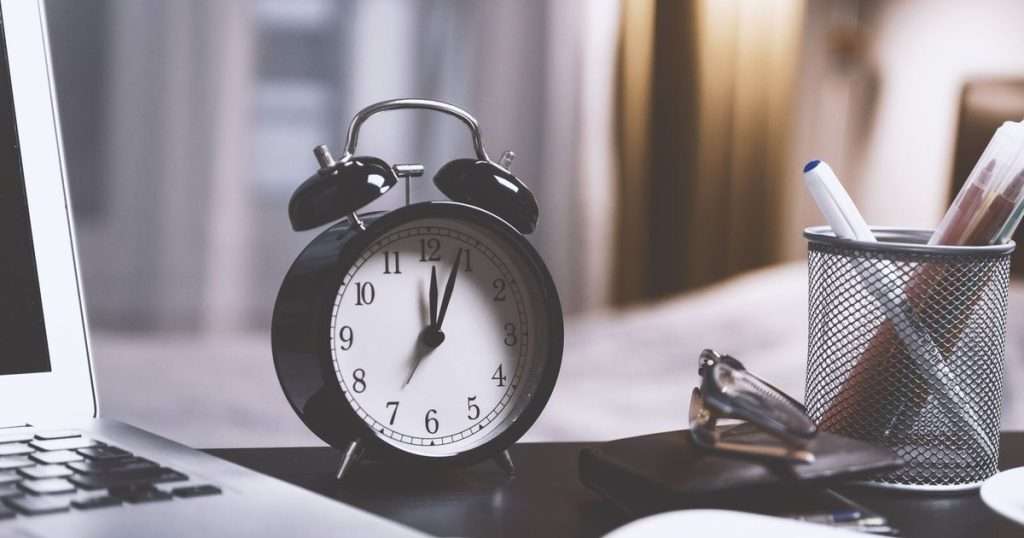 Programar un correo en Outlook: reloj despertador negro de campanas sobre un escritorio, junto a un portátil, unas gafas y un lapicero