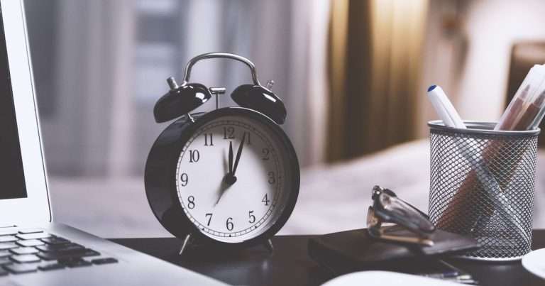 Programar un correo en Outlook: reloj despertador negro de campanas sobre un escritorio, junto a un portátil, unas gafas y un lapicero