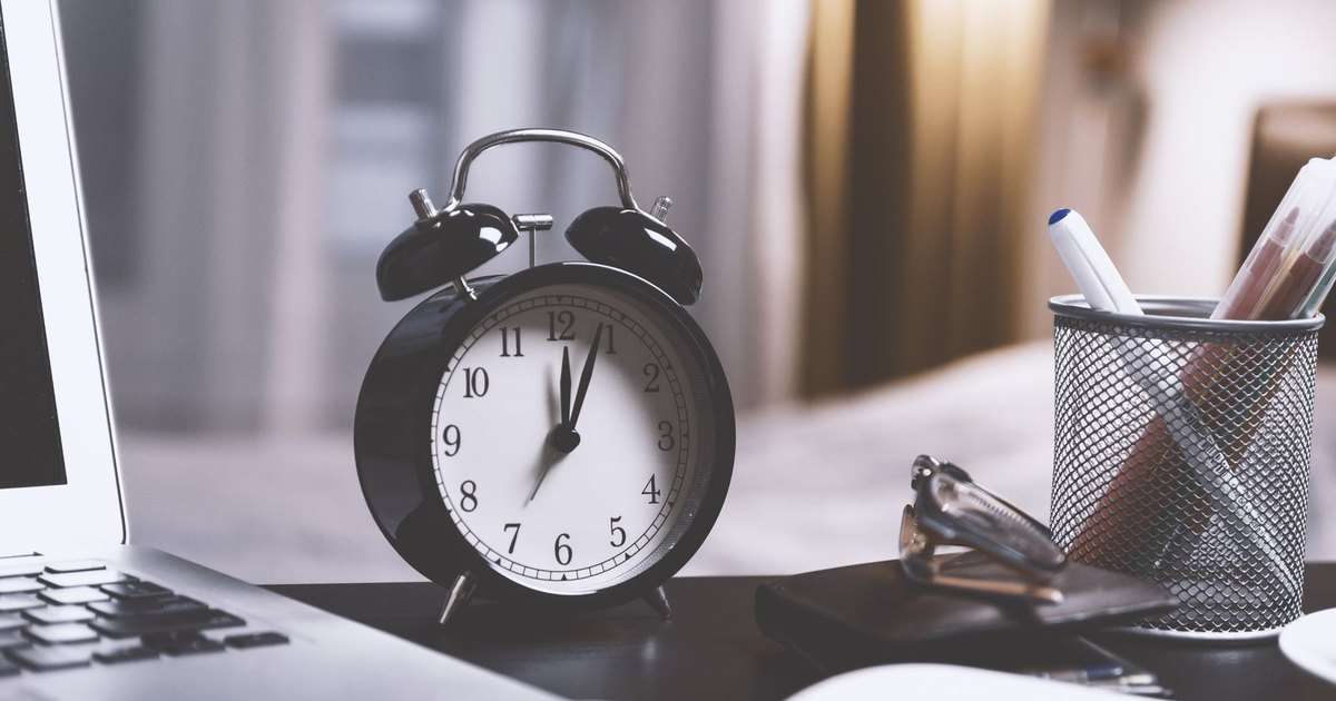 Programar un correo en Outlook: reloj despertador negro de campanas sobre un escritorio, junto a un portátil, unas gafas y un lapicero