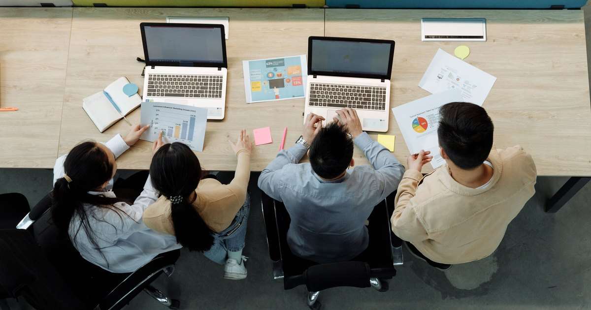 Qué es SharePoint: cuatro compañeros vistos desde arriba, de espaldas, trabajando juntos en una mesa compartida con dos portátiles, gráficos impresos, notas adhesivas y una libreta