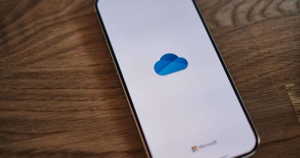 Sincronizar OneDrive: teléfono sobre una mesa de madera con la aplicación de OneDrive abriéndose, con el logotipo de la nube azul y la marca Microsoft en la pantalla